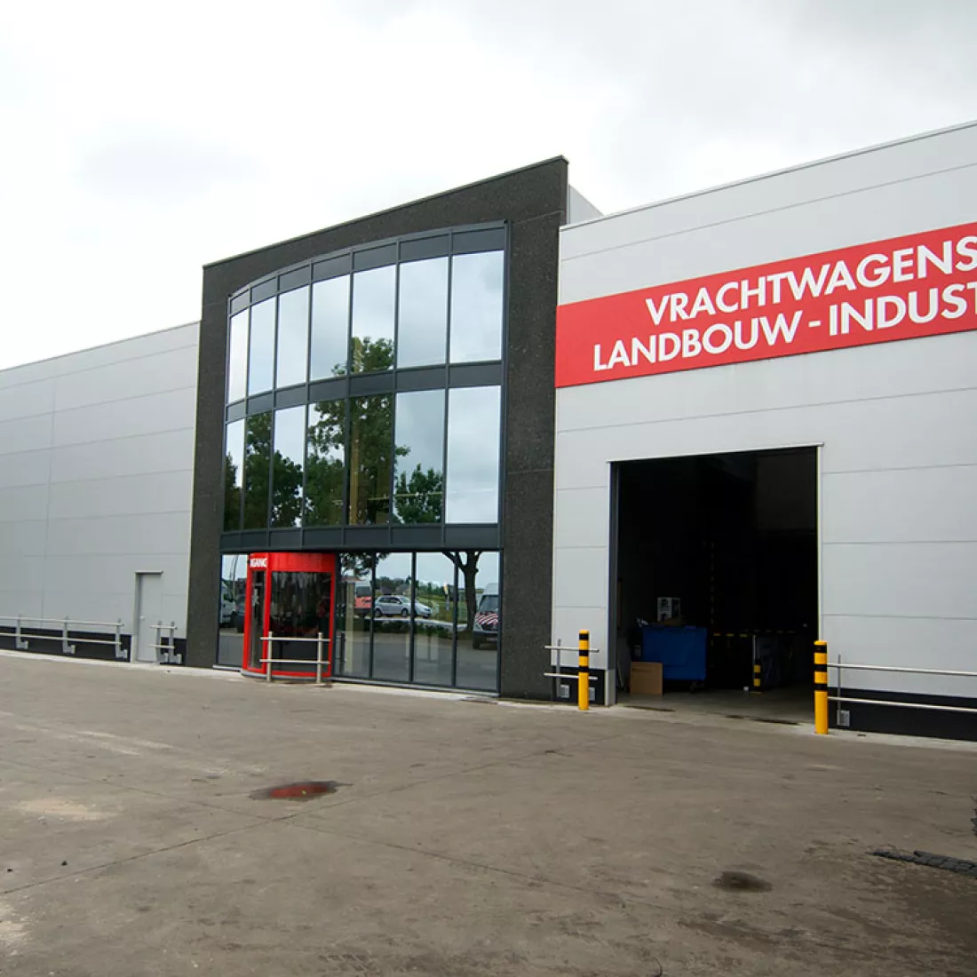 Vooraanzicht TSC. Het gebouw is opgebouwd met een combinatie van materialen. Links bestaat het gebouw uit grijze platen. In het midden bevindt zich de inkom. Dit deel van het gebouw is zwart en bevat veel ramen die bestaan uit spiegelglas en drie verdiepingen. De magazijnen rechts en links van de inkom bestaan uit grijze platen. Rechts bevindt zich een poort. Boven de poort hangt een rode rechthoek. Hierop staat in witte letters "Vrachtwagens landbouw - industrie".