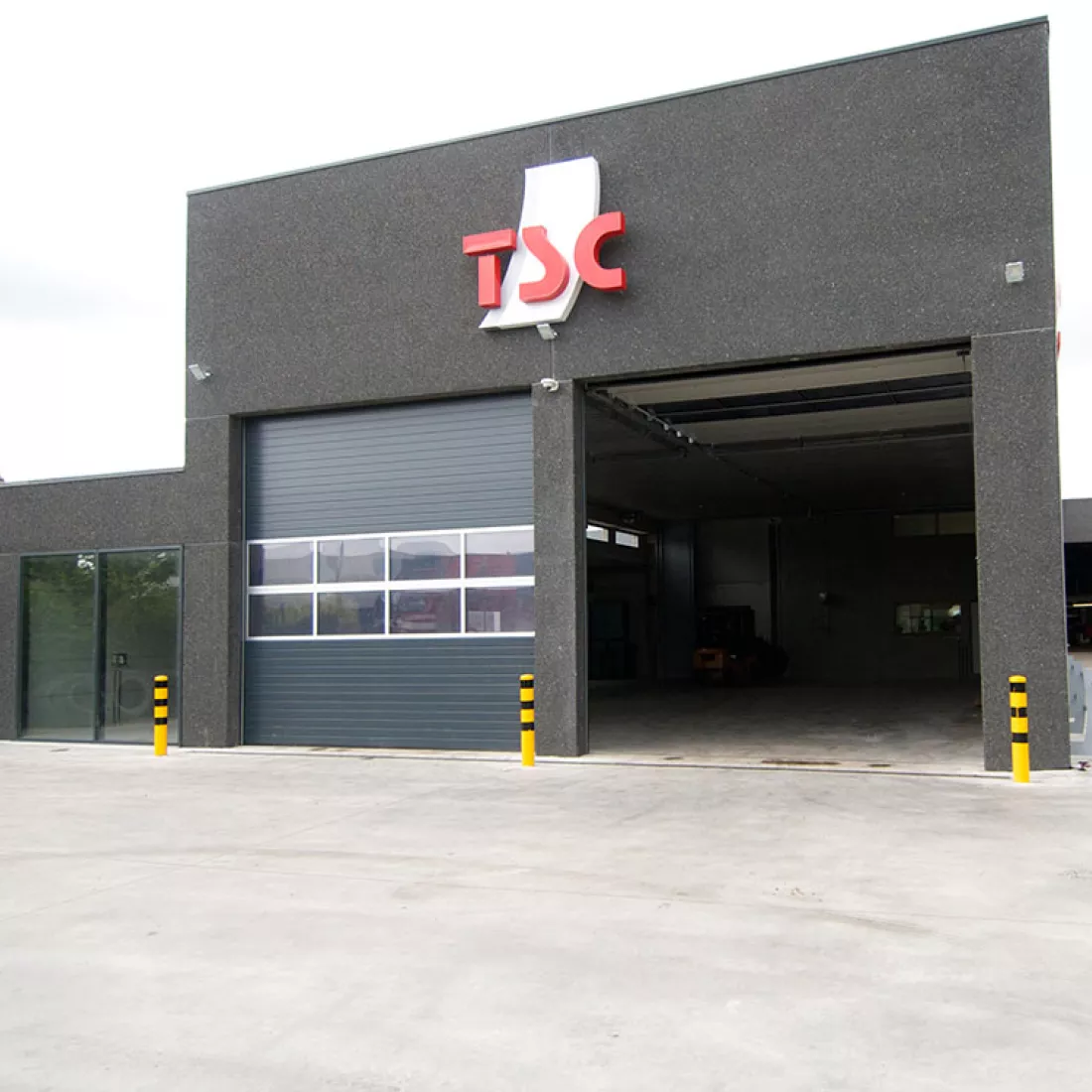 Vooraanzicht opslaghal banden - TSC. Bovenaan het gebouw hangt het logo van TSC Het gebouw is zwart en bevat twee zwarte poorten.