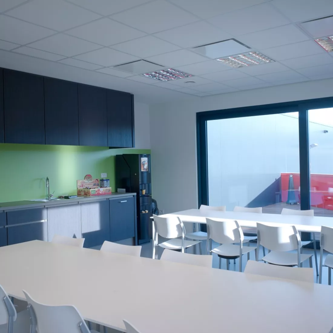 Binnenkant kantoor - DIBO. Het is een afbeelding van de lunchruimte. Links bevindt zich een keuken met een groene wand. De keuken is zwart. Rechts van de keuken staan witte tafels met witte stoelen. Achteraan bevindt zich een schuifraam.