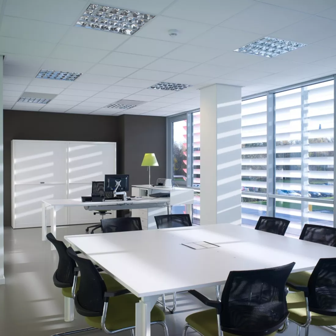 Binnenkant kantoor - DIBO. Het is een afbeelding van de vergaderruimte. Rechts bevinden zich grote ramen. De muur achteraan is bruin en er staat een witte kast tegen. Voor de kast staat een bureau. Voor de bureau bevindt zich een vierkante vergadertafel met acht stoelen rond.