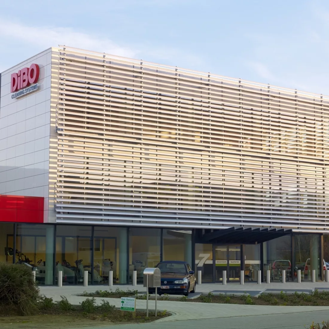 Vooraanzicht bedrijfspand met kantoor - DIBO Cleaning Systems. Het gebouw bestaat uit witte en rode platen. Links hangt het logo van DIBO. Voor het gebouw zijn parkeerplaatsen voorzien.