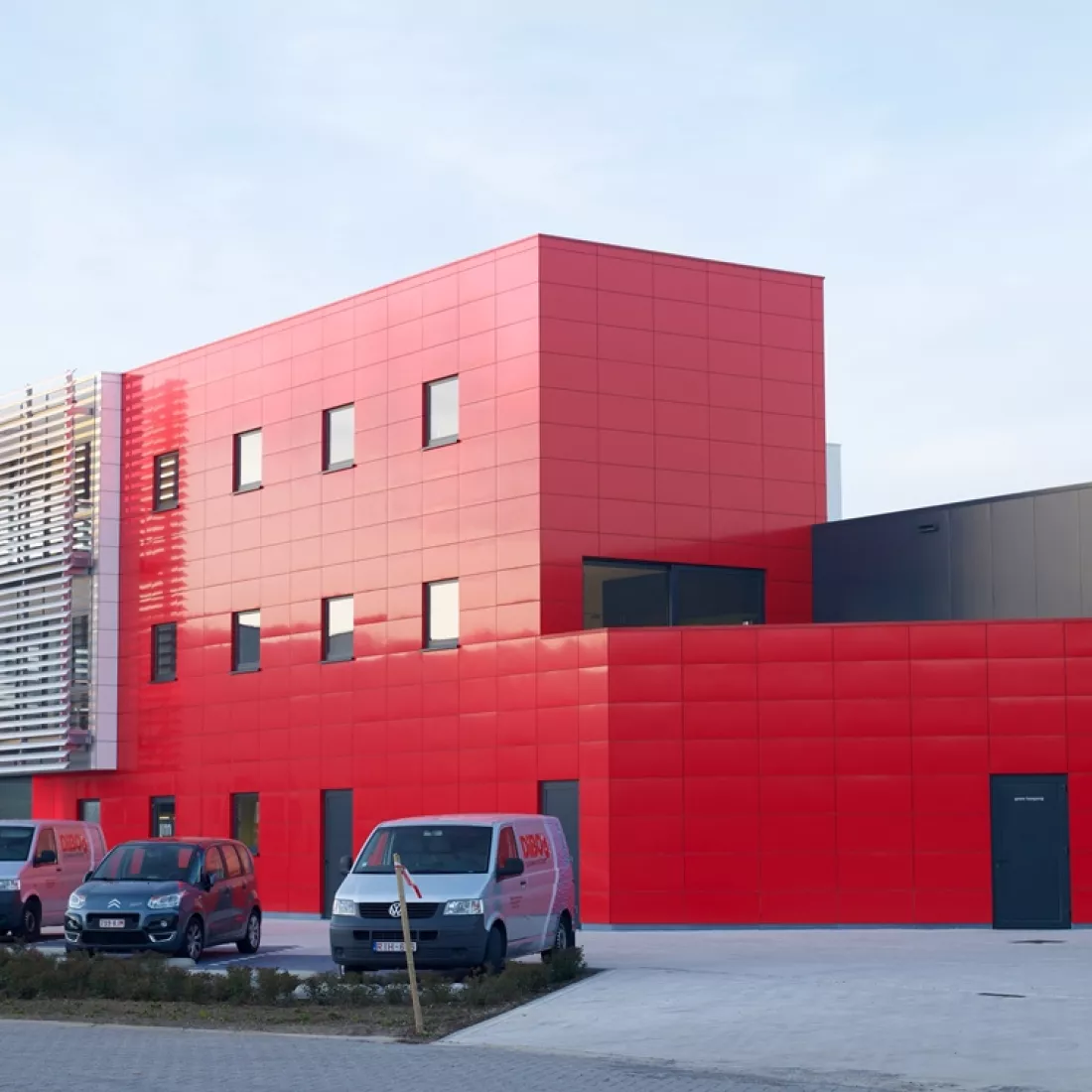 Zijaanzicht bedrijfspand met kantoor - DIBO Cleaning Systems. Het gebouw bestaat uit rode platen en bevat zwarte deuren. Langs de zijkant heeft het gebouw kleinere ramen. Het gebouw bestaat uit drie verdiepingen.