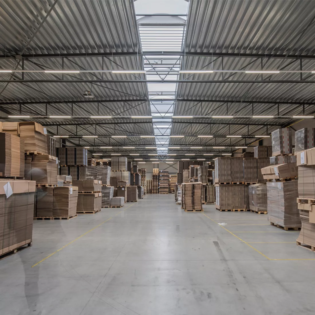 Binnenkant magazijn Corpack met in het plafond een lichtstraat. Op de grond zijn afgebakende vakken aan de hand van gele lijnen. In de vakken staan materialen opgeslagen.
