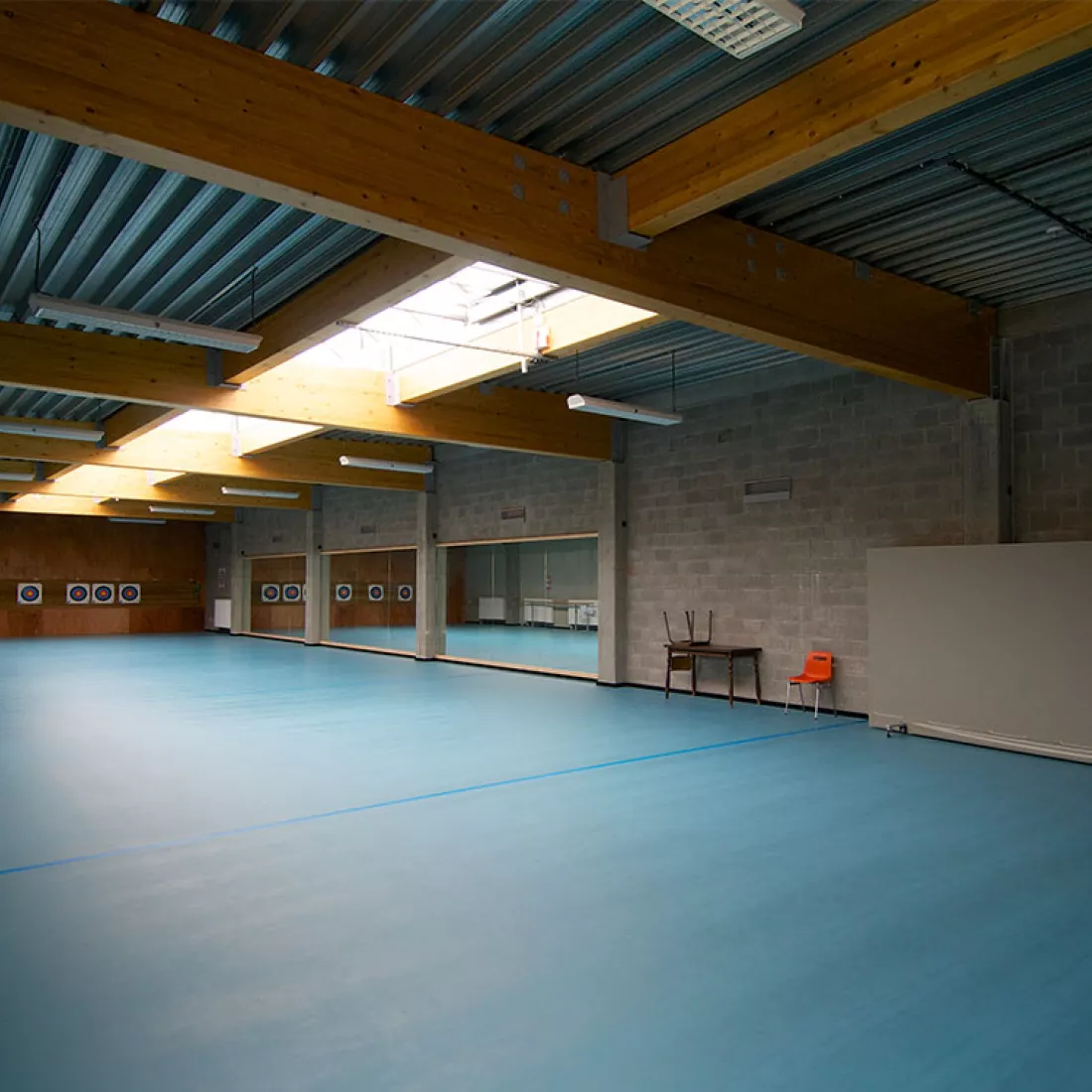 DCA Sportcomplex - binnenkant. Het plafond bevat een lichtstraat. Rechts achteraan hangen er spiegels tegen de muur. Achteraan links hangen boogschietdoelen. De vloer is blauw.