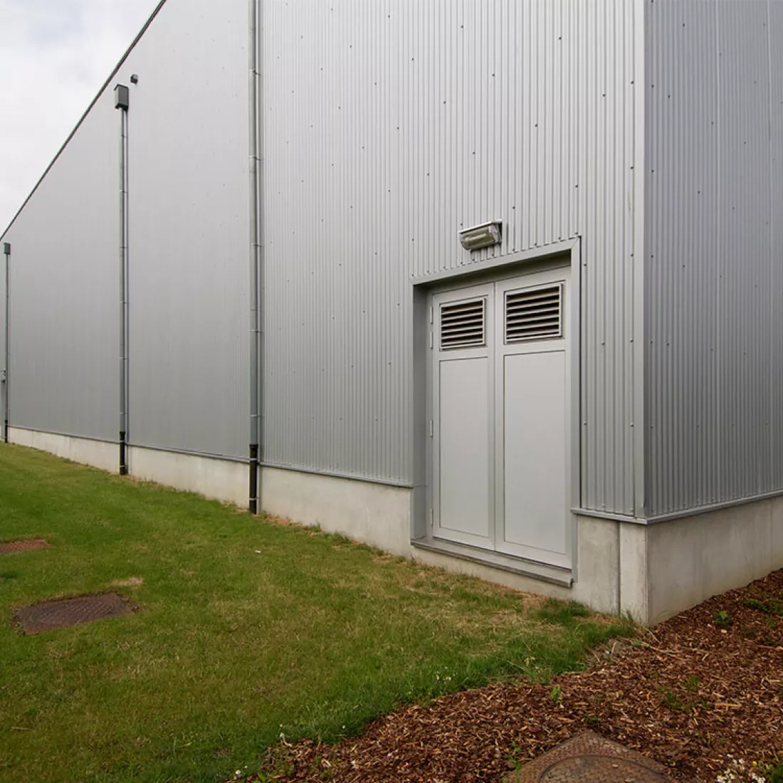 DCA Sportcomplex - buitenkant. Het gebouw is opgebouwd met een combinatie van materialen zoals beton. Links naast het gebouw bevindt zich een grasveld. Het gebouw bevat een grijze deur.