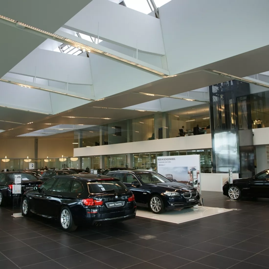 Binnenkant Centrauto - showroom. Het gebouw bestaat uit twee verdiepingen. Op de beneden verdieping staan er verschillende auto's tentoongesteld. Achter de auto's bevindt zich een glazenwand. Op de bovenste verdieping bevinden zich kantoren. Er bevindt zich ook een lift.