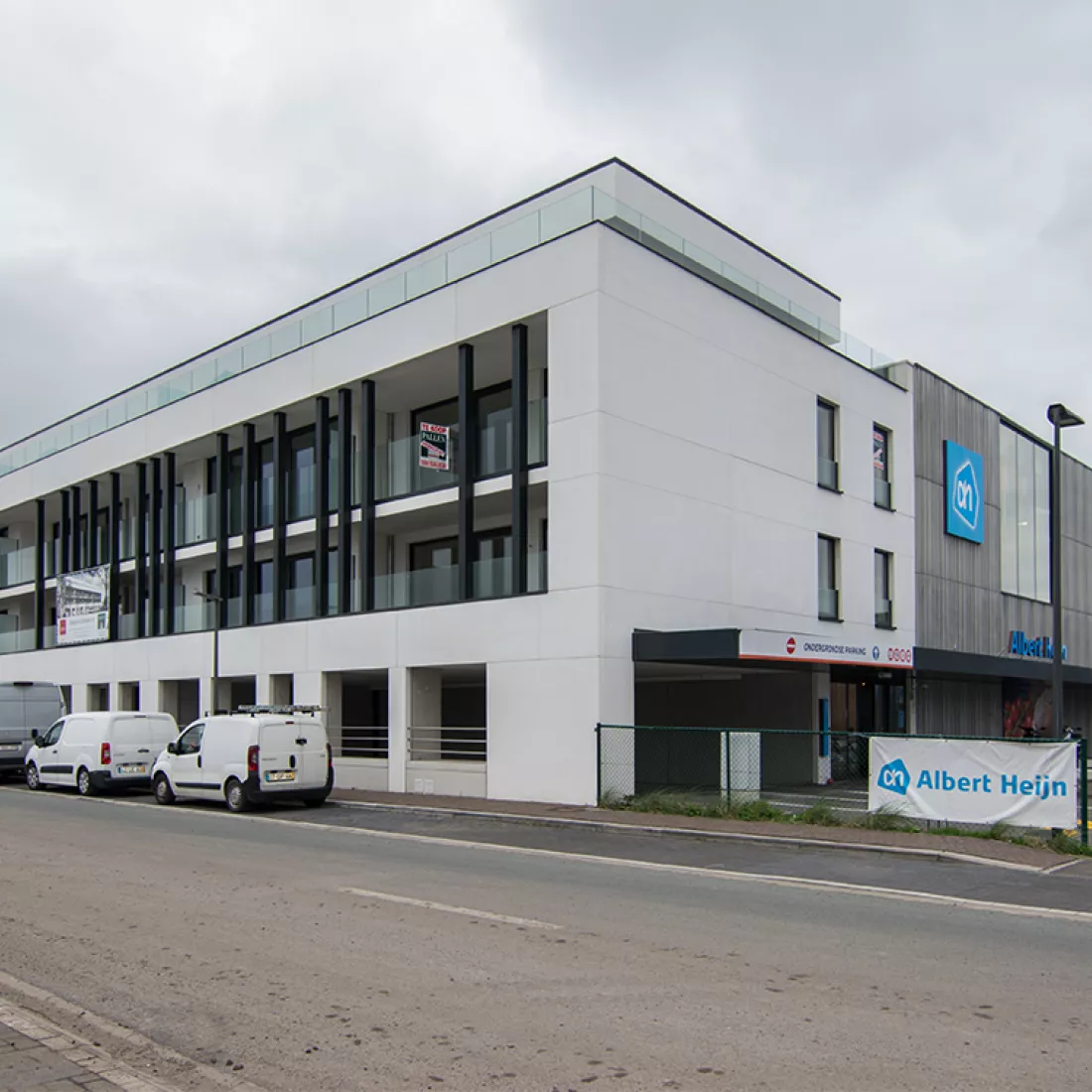 Zijaanzicht Duin & Water shops. Zijaanzicht Duin & Water shops. De gelijkvloers is een parking. Het is een wit gebouw. Achter het gebouw bevindt zich rechts een Albert Heijn.