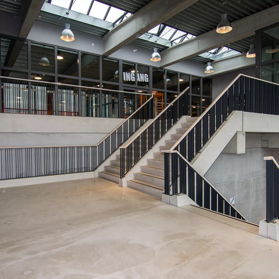 Trappenhal Duin & Water shops. Trappenhal Duin & Water shops. De afbeelding is genomen vanop de trap. Deze trap gaat naar de inkom van de Basic Fit. Het is een betonnen trap met een zwarte railing. Boven de trap is een plafond. In het plafond bevindt zich een lichtstraat.