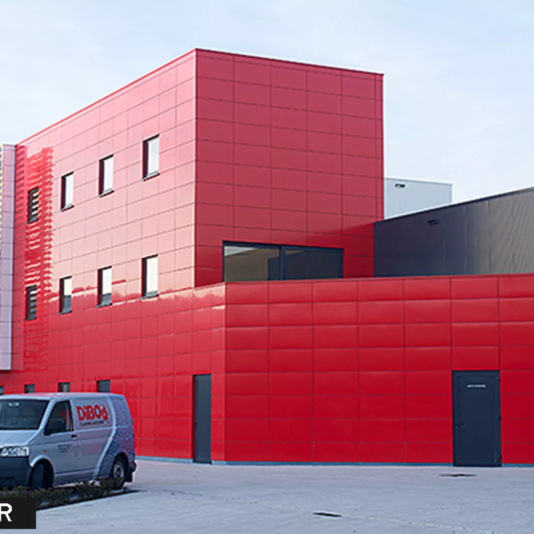 Zijaanzicht werkgebouw DIBO voor renovatie gevel. Het gebouw bevat rode tegels en ramen met zwarte omranding. De deuren zijn ook zwart.