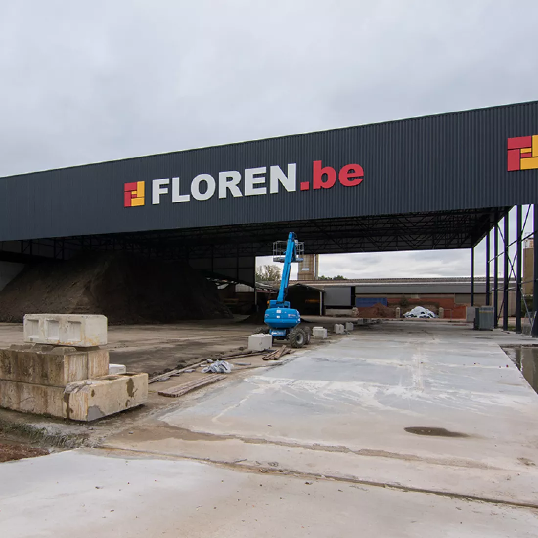 Vooraanzicht luifel kleihal Floren. De luifel is zwart en bevat het logo van Floren. Tijdens de opbouw is staal gebruikt. Voor de luifel staat een blauwe hoogwerker.