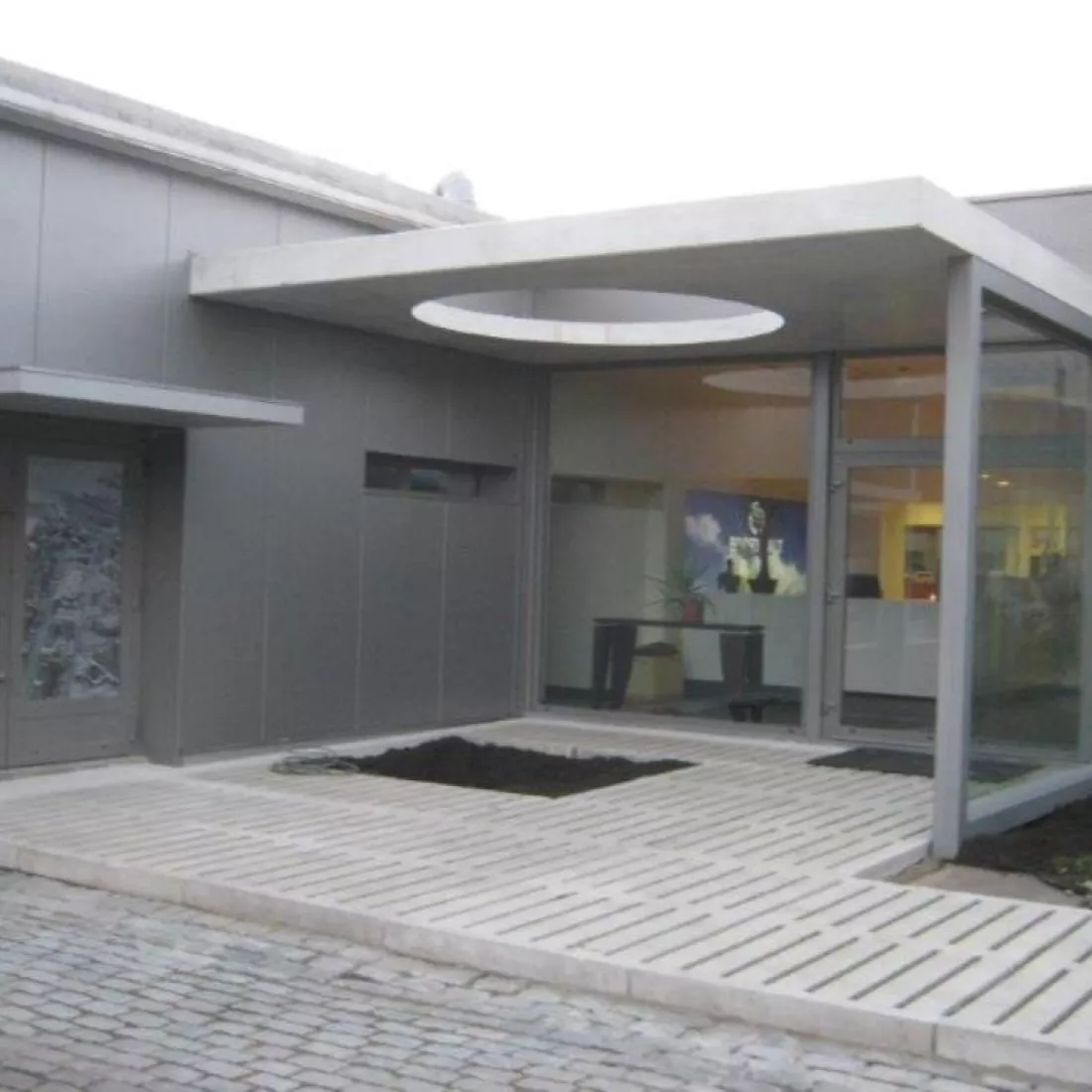 Zijaanzicht Boortmalt inkom. Het gebouw is grijs en bevat grote ramen. Tegen het gebouw hangt een luifel met een gat erin. Het gat is een cirkel voor een boom er door te laten groeien.
