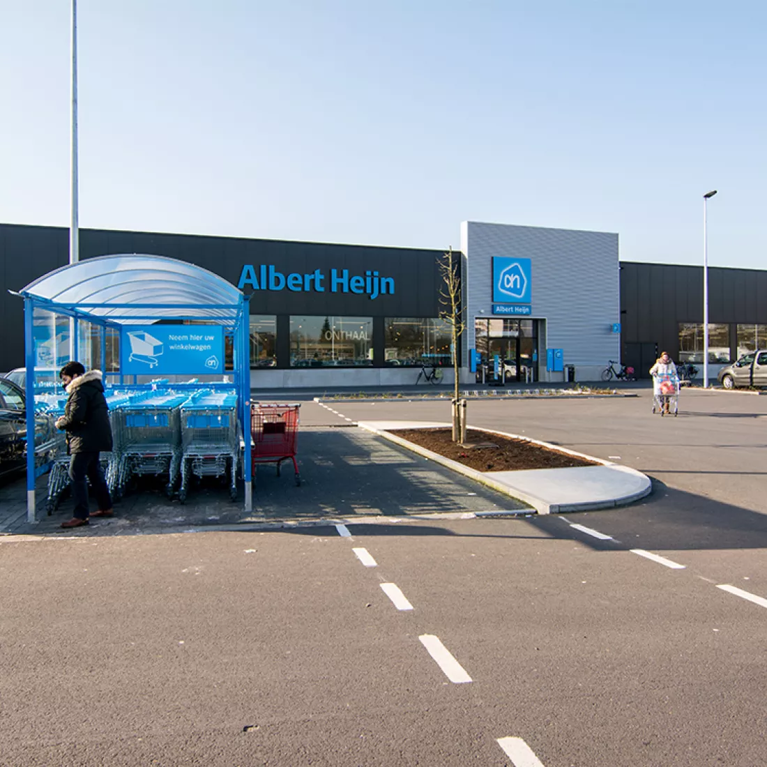 Handelsruimten Immo Euro - Albert Heijn. Het betonnen gebouw bestaat uit zwarte en grijze platen. Het logo en de letters van Albert Heijn hangen bovenaan het gebouw. Op de parking is een plek voor winkelkarren voorzien.
