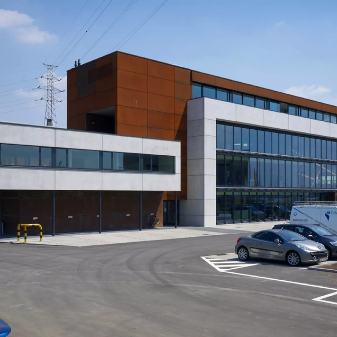 Zijaanzicht Ivens kantoor. Het kantoor bestaat uit veel ramen. Het gebouw bevat witte en bruine platen. Het kantoor bestaat uit vier verdiepingen. Voor het gebouw is een parking.