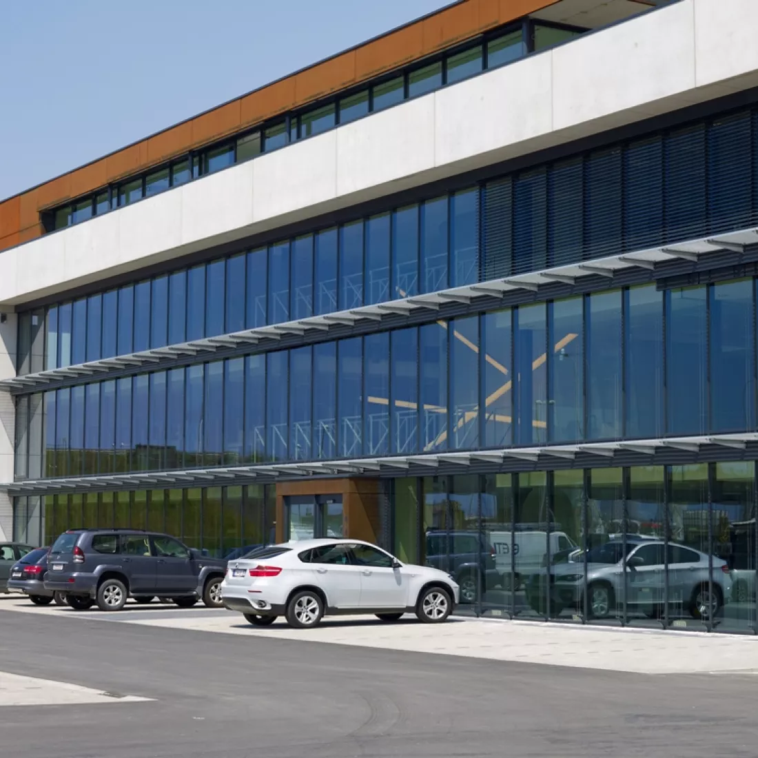 Ivens kantoor. Voor het kantoor bevindt zich een parking. Het gebouw bestaat uit vier verdiepingen en veel ramen.