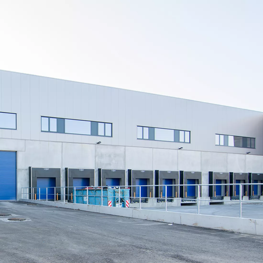 Buitenkant magazijn - Keetberg Logistics Center. Het magazijn bevat een dock of laadruimte, dit is voor het laden en lossen van vrachtwagens. De poorten zijn blauw. Links naast het dock bevindt zich een grotere blauwe poort. Het gebouw bestaat uit twee verdiepingen. Het tweede verdiep bevat ramen.
