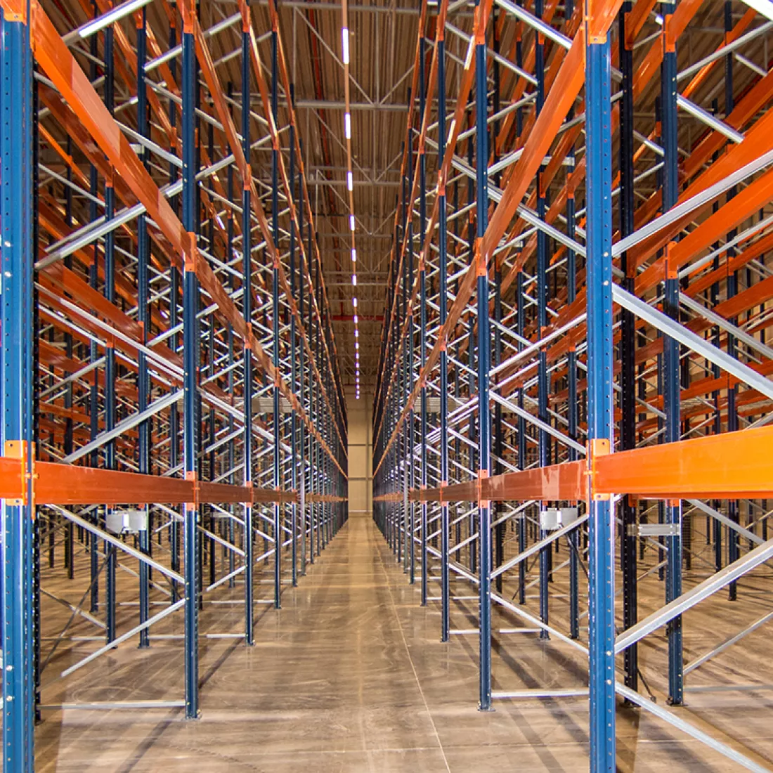 Binnenkant magazijn - Keetberg Logistics Center. In het magazijn staan gangen met rekken. De rekken zijn blauw met oranje. In deze rekken kunnen producten worden opgeslagen.
