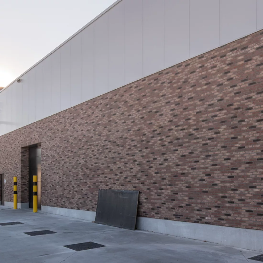 Zijaanzicht nieuwbouw magazijn Nauta Holding. De onderkant van het magazijn bestaat uit rode steen en de bovenkant uit grijze wanden. Links bevindt zich een zwarte poort en een zwarte deur.