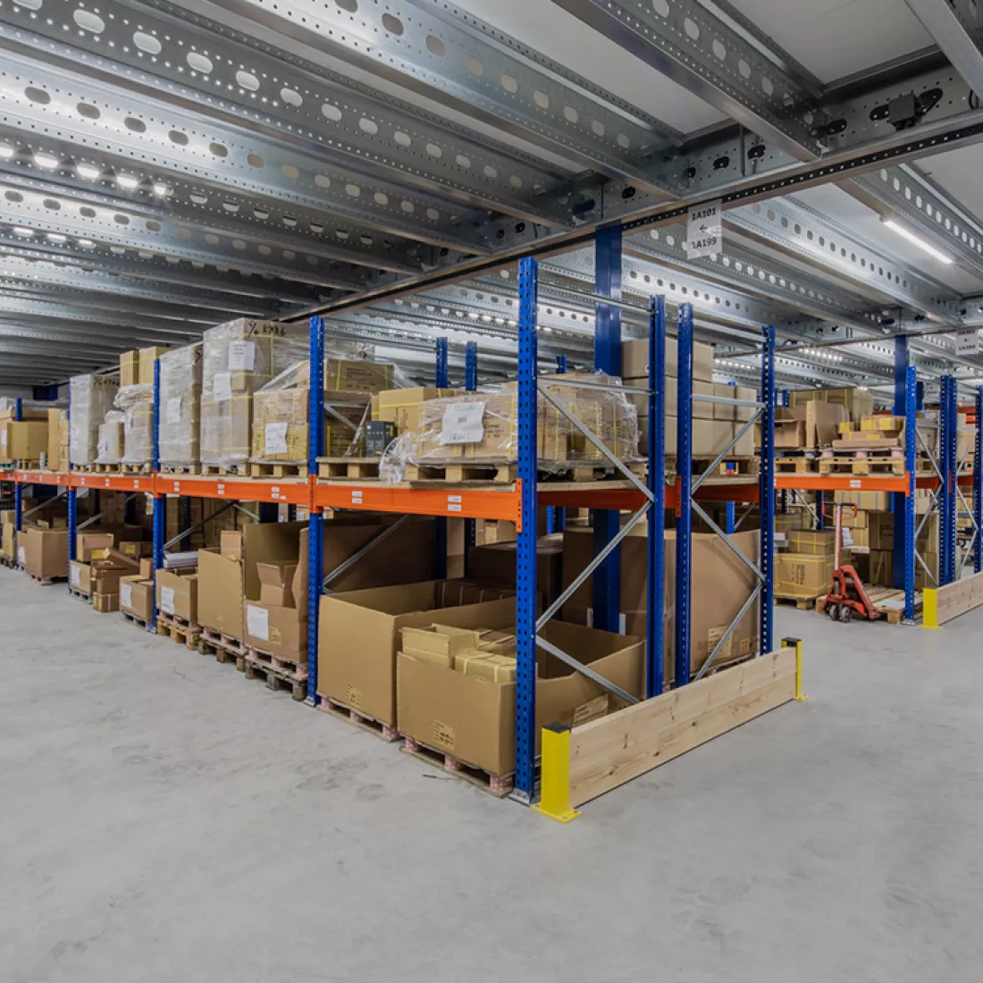 Binnenkant nieuwbouw magazijn Nauta Holding. In het magazijn staan rijen met oranje en blauwe rekken. In de rekken staan kartonnen dozen opgeslagen. Deze doezen staan op paletten.