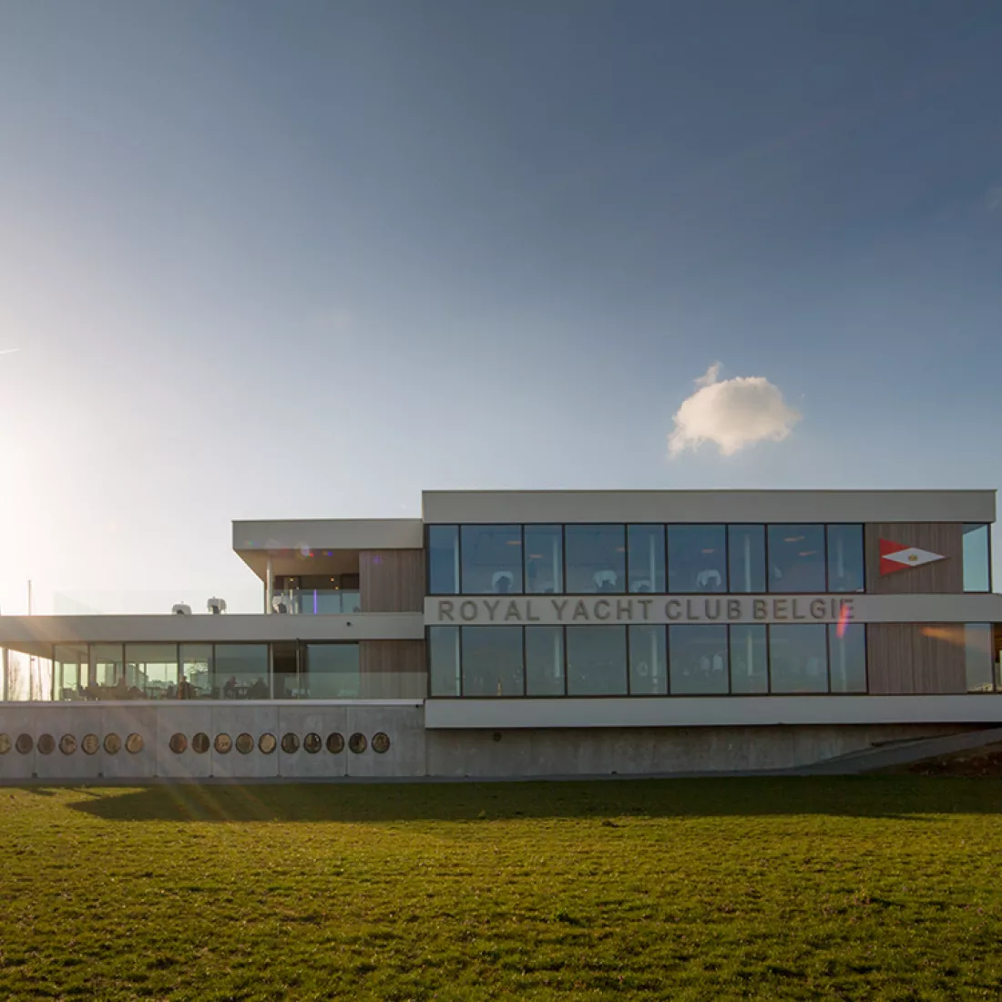 Vooraanzicht Royal Yacht Club België. Het gebouw is opgebouwd met beton. Het heeft drie verdiepingen en veel ramen. Voor het gebouw bevindt zich een grasveld. 