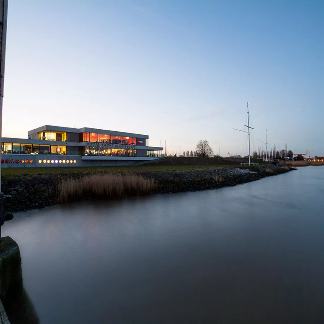 Vooraanzicht Royal Yacht Club België. Het gebouw is opgebouwd met beton. Het heeft drie verdiepingen en veel ramen. Voor het gebouw bevindt zich een grasveld en de Schelde.