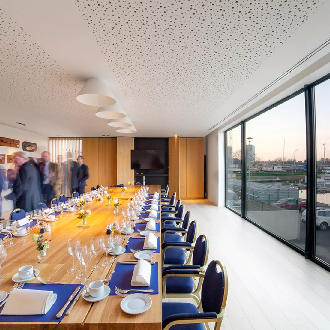 Binnenkant Royal Yacht Club België. In het midden staat een rechthoekige houtentafel. De tafel is mooi opgedekt. Rond de tafel staan blauwe stoelen. Rechts bevindt zich een groot raam. Tegen de achterste muur hangt een TV.