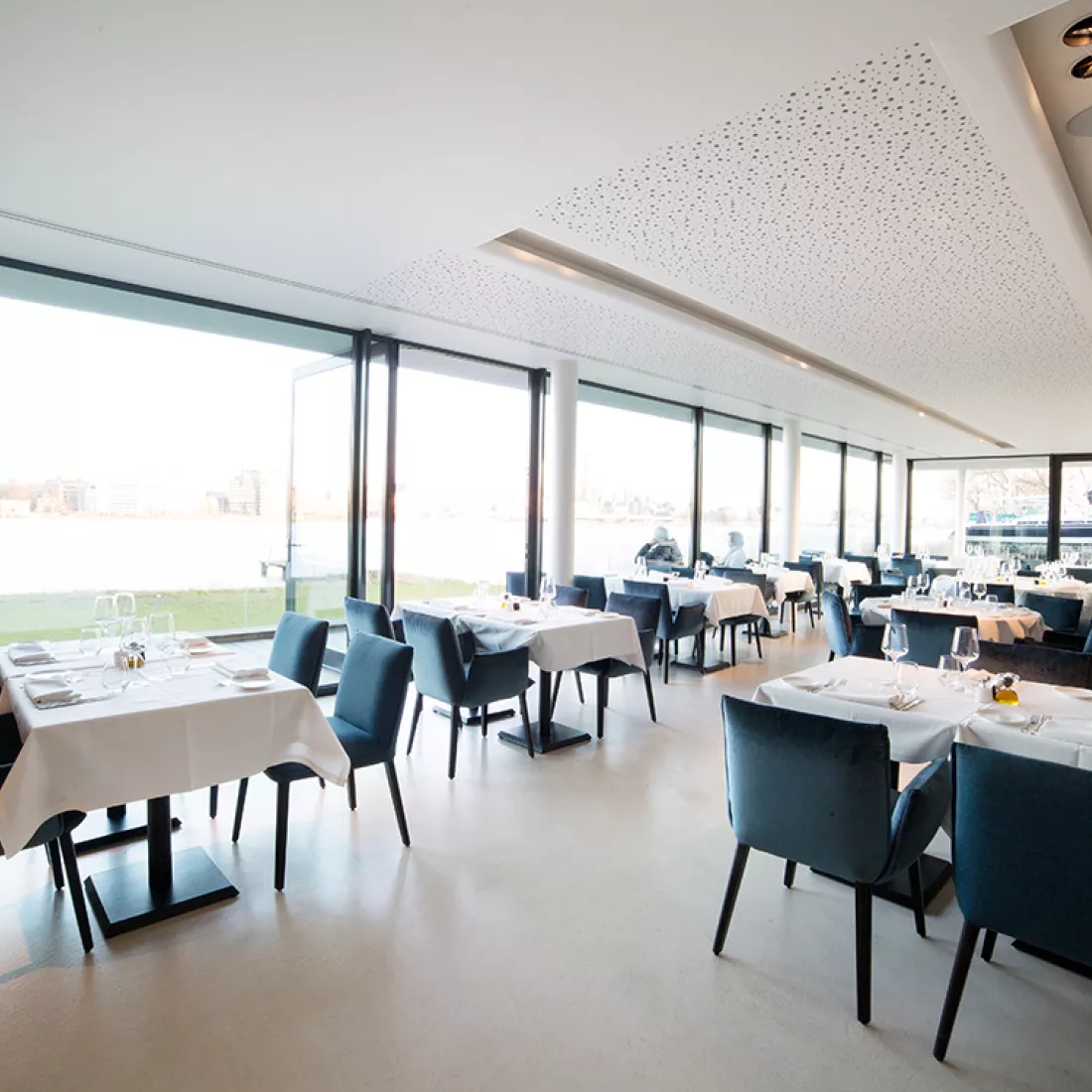 Binnenkant restaurant Royal Yacht Club België. Er bevinden zich mooi opgedekte tafels met blauwe stoelen rond. Links bestaat de muur uit ramen. Het plafond en de vloer is wit.
