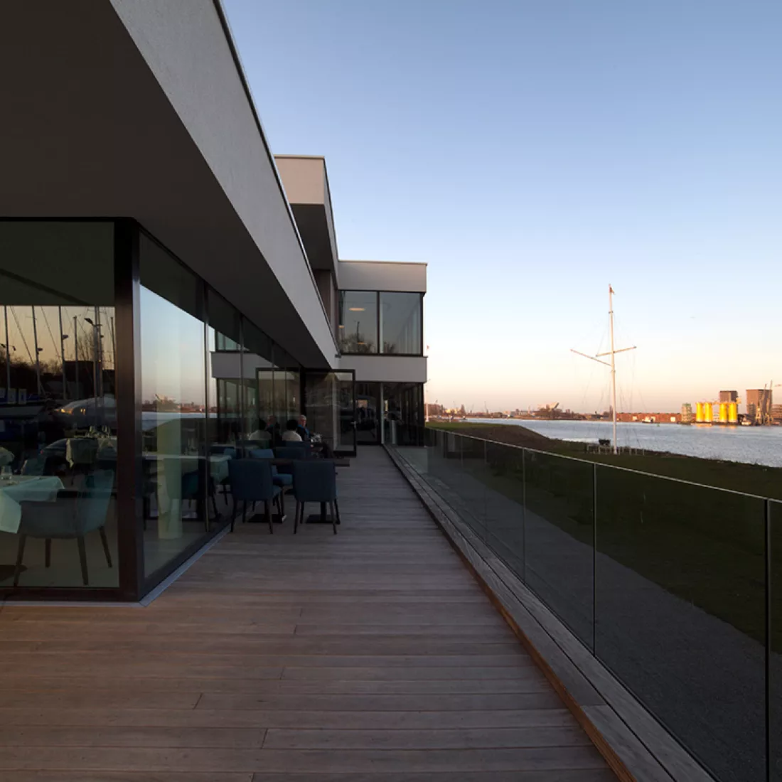 Balkon restaurant Royal Yacht Club België. Rechts bevindt zich de Schelde. Links het restaurant. Er staan enkele tafels buiten op het balkon. Het balkon heeft een glazen railing.
