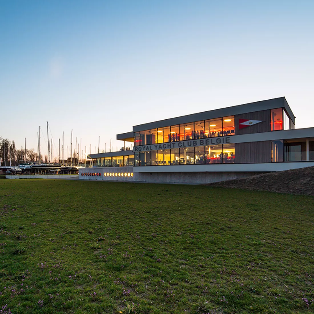 Vooraanzicht Royal Yacht Club België. Het gebouw is opgebouwd met beton en heeft drie verdiepingen. Links naast het gebouw bevinden zich boten. Voor het gebouw bevindt zich een grasveld.