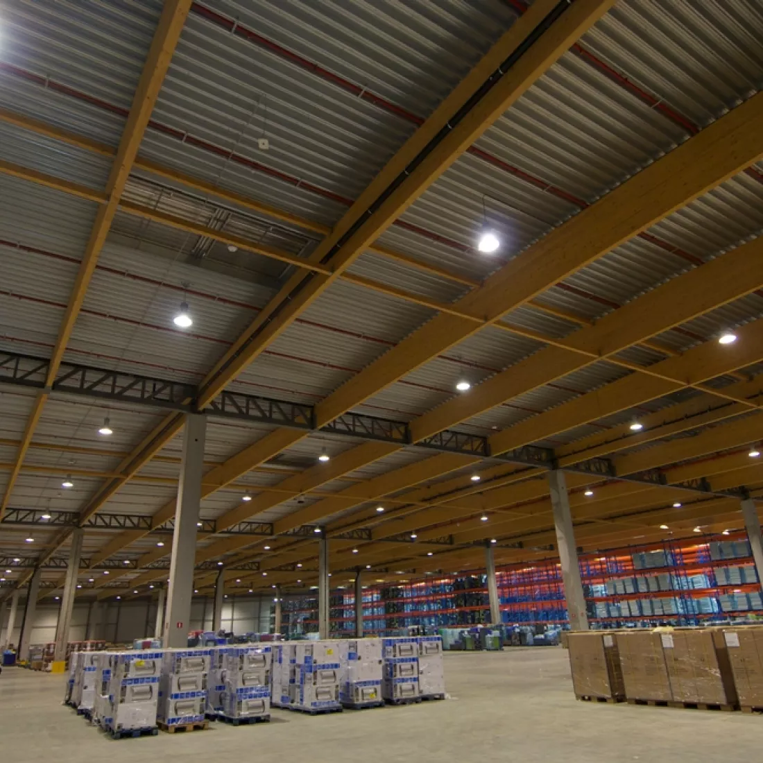 Binnenkant distributiecentrum ASL & ASL. Er is een combinatie van materialen gebruikt zoals hout bij het plafond en betonpilaren. Er staan verschillende materialen opgeslagen. Achteraan staan de materialen in oranje met blauwe rekken opgeslagen.