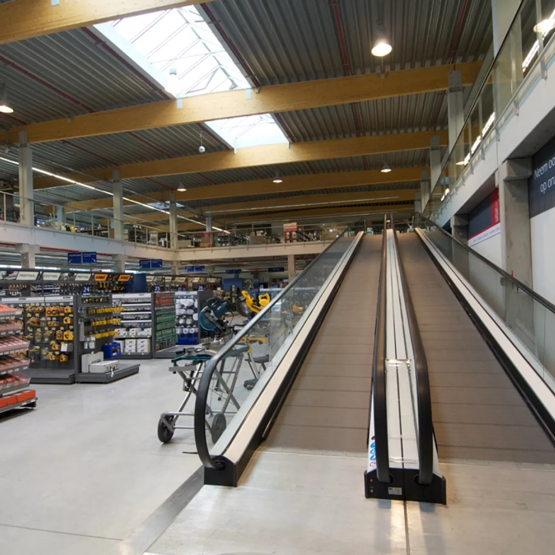 Binnenkant winkel METALeuven. Rechts bevinden zich rolpaden omhoog naar de tweede verdieping. Die verdieping heeft een glazen railing. Links bevinden zich rekken waar materialen tentoongesteld liggen. Het plafond bevat een lichtstraat.