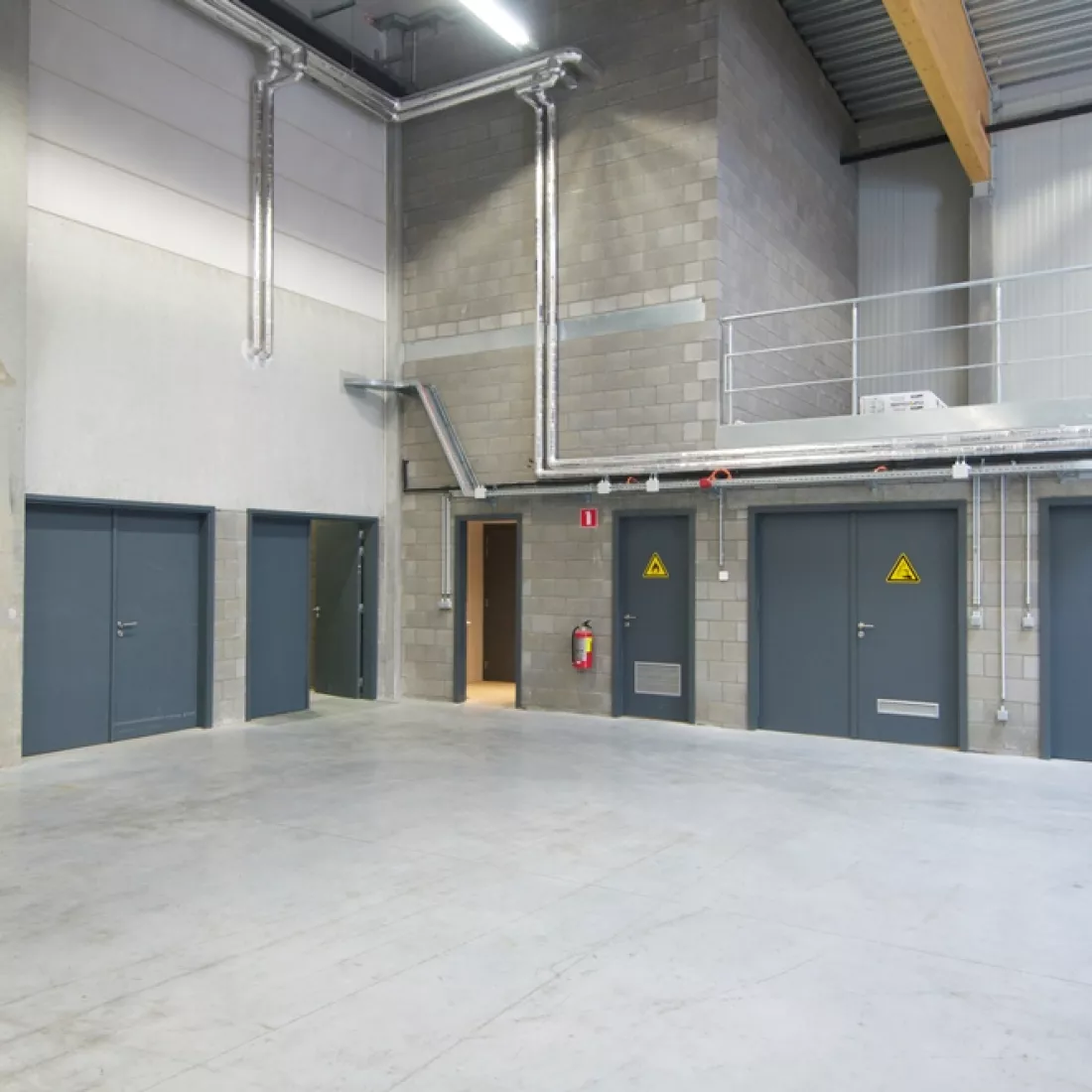 Binnenkant werkplaats METALeuven. Het gebouw is opgebouwd met een combinatie van materialen. Links en achteraan bevinden zich muren met grijze deuren in.