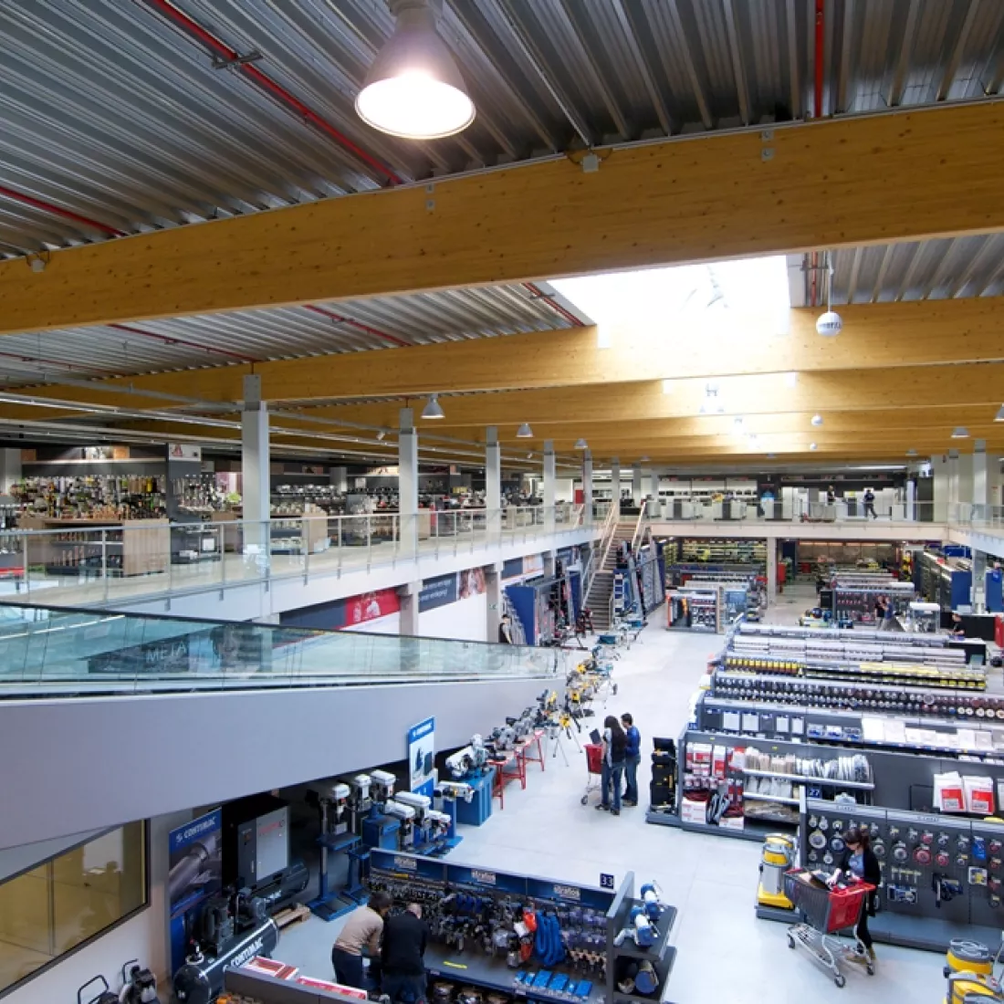 Binnenkant winkel METALeuven. De afbeelding is vanaf de tweede verdieping gemaakt. Links bevinden de rolpaden zich en links achteraan bevinden zich trappen. De verdieping heeft een glazen railing. De producten staan opgeslagen in rekken.