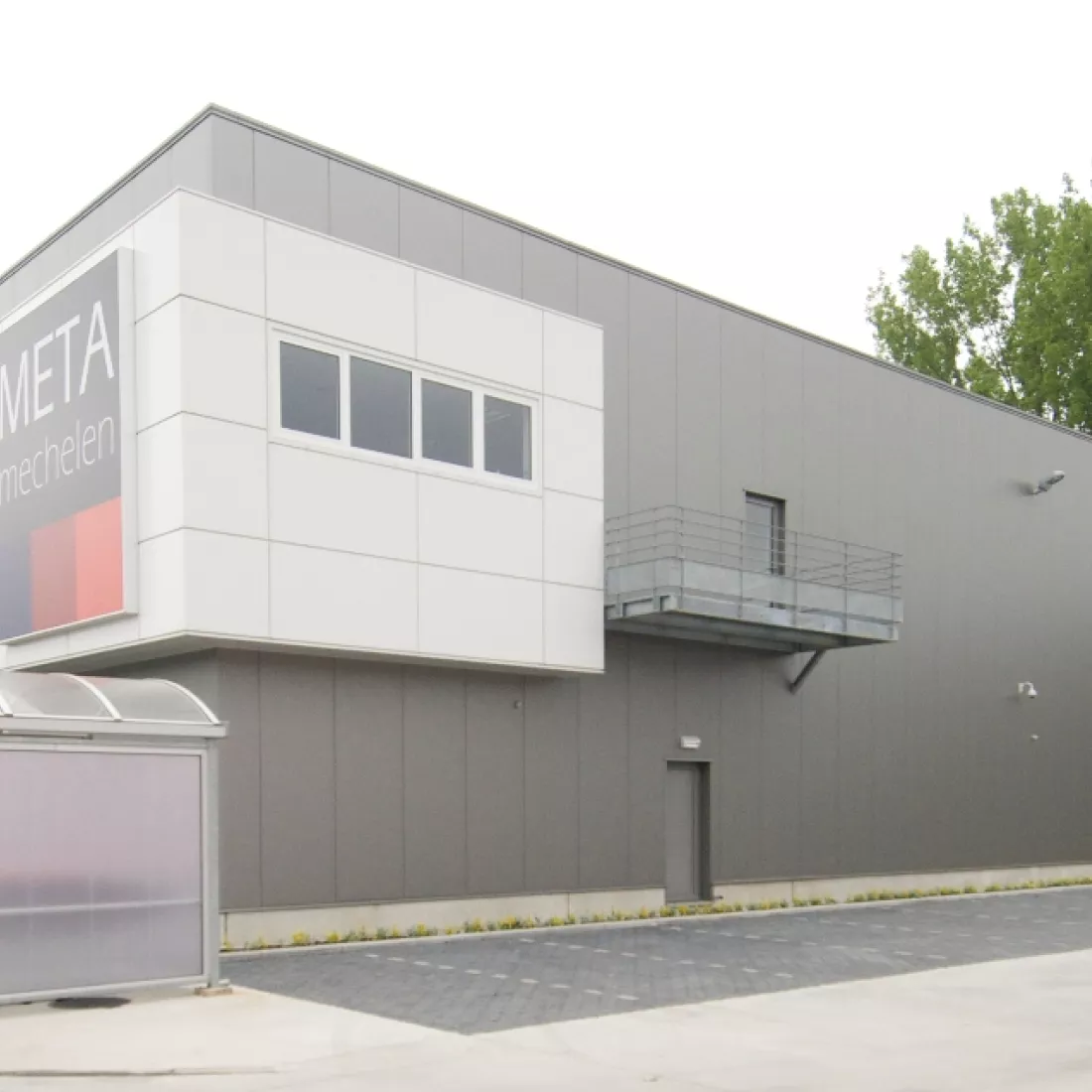 Zijaanzicht METALeuven. Rechts naast het gebouw zijn ook parkeerplaatsen voorzien. Het gebouw bestaat uit grijze en witte platen. Het gebouw bevat grijze deuren.