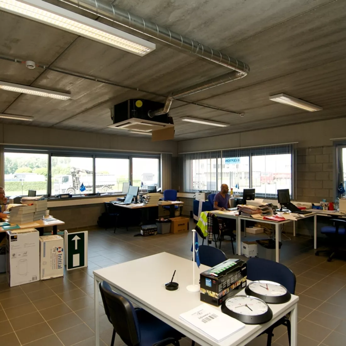 Binnenkant kantoor distributiecentrum ASL & ASL. Het kantoor is opgebouwd met beton. Er staan witte tafels met blauwe stoelen aan. De buitenmuren bevatten ramen.
