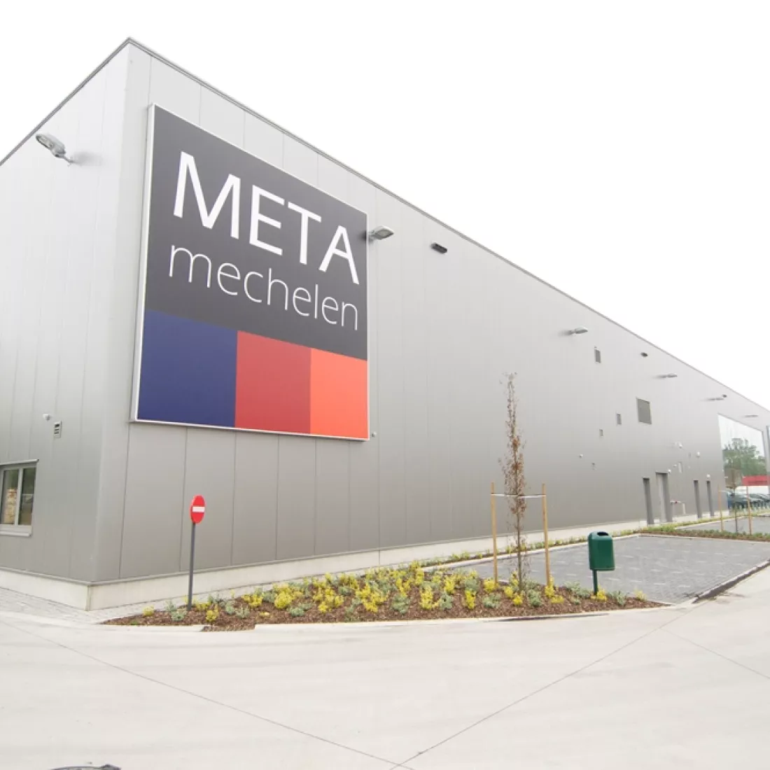 Zijaanzicht METALeuven. Voor het gebouw zijn parkeerplaatsen voorzien. Links bevindt zich het gebouw. Het gebouw bestaat uit grijze platen. Links hangt het logo van META.