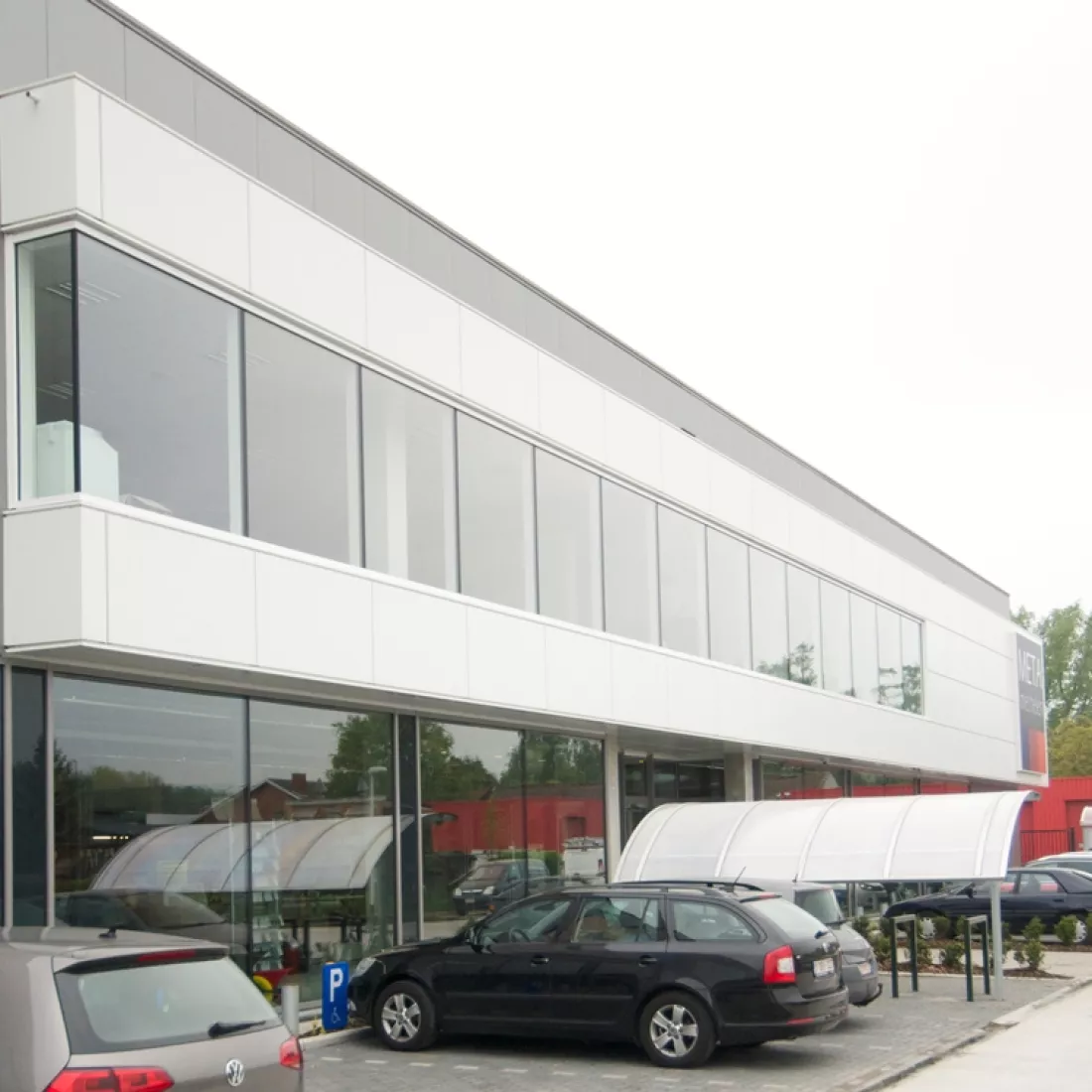 Kantoor en inkom METALeuven. Het kantoor en de inkom bestaat uit witte platen en bevat grote ramen. Voor het gebouw zijn parkeerplaatsen voorzien.