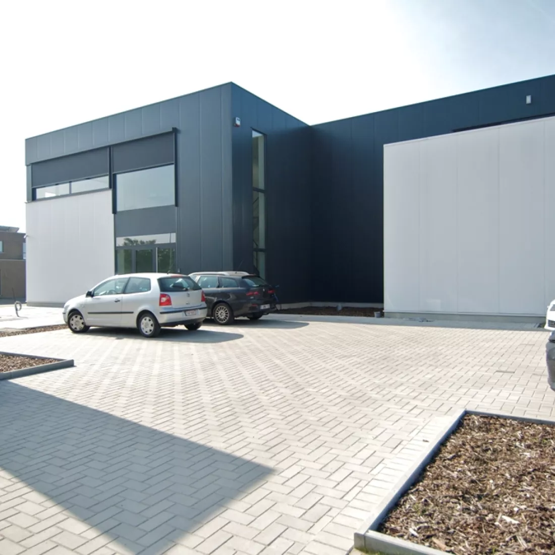 Vooraanzicht productiehal en kantoor Argina. Het gebouw is wit met zwart en bevat enkele ramen. Voor het gebouw zijn parkeerplaatsen voorzien.