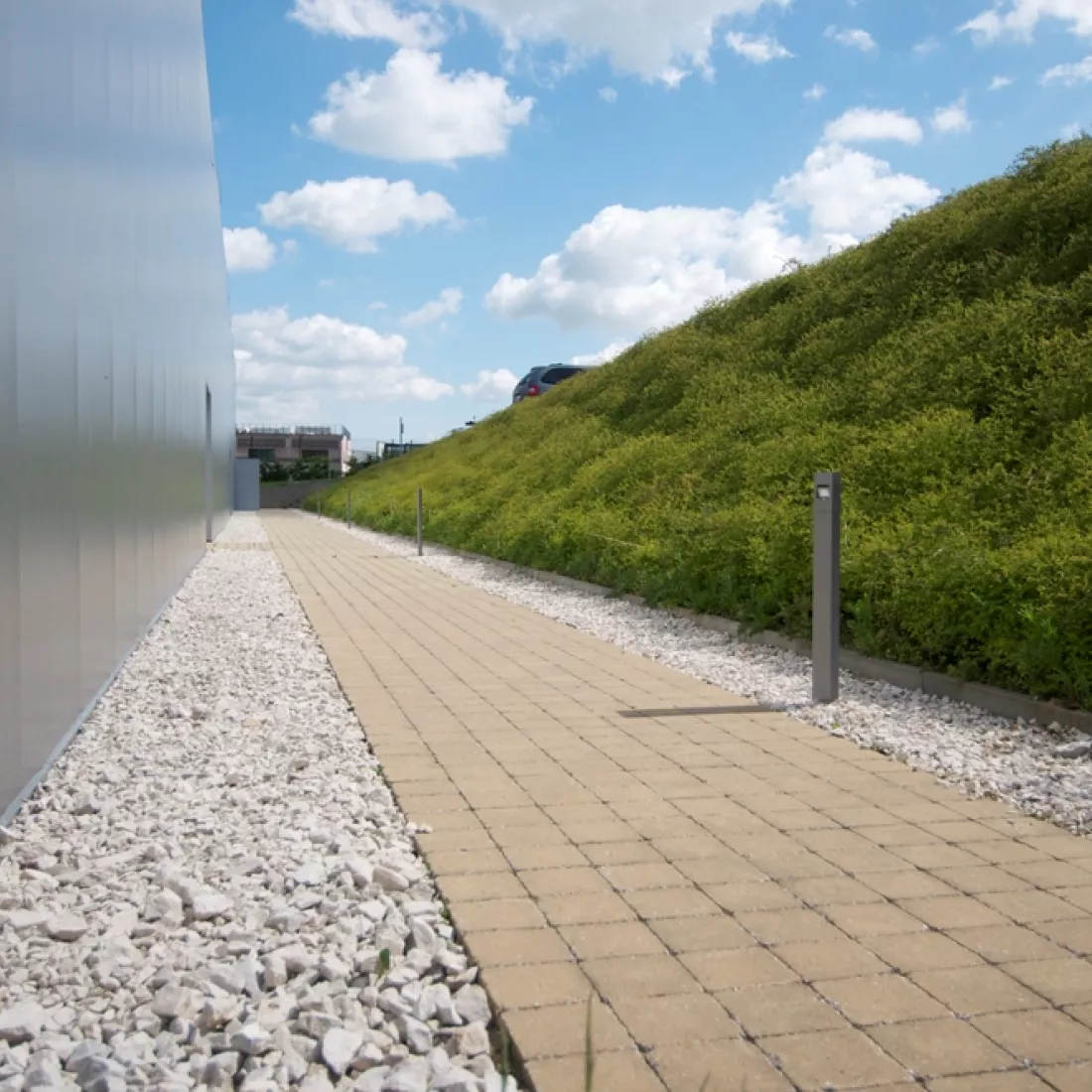 Zijaanzicht kantoor JNL. Links bevindt zicht het grijze kantoor. Naast het kantoor liggen steentjes, daarna een pad dat bestaat uit vierkante stenen en langs de andere kant weer steentjes. Dan is er een helling dat bestaat uit groene planten.