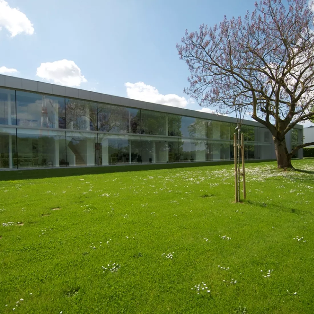 Buitenkant kantoor JNL. Voor het kantoor is een groot grasveld. Het kantoor bestaat uit heel veel ramen. Het is een grijs gebouw en gebouwd met staal.