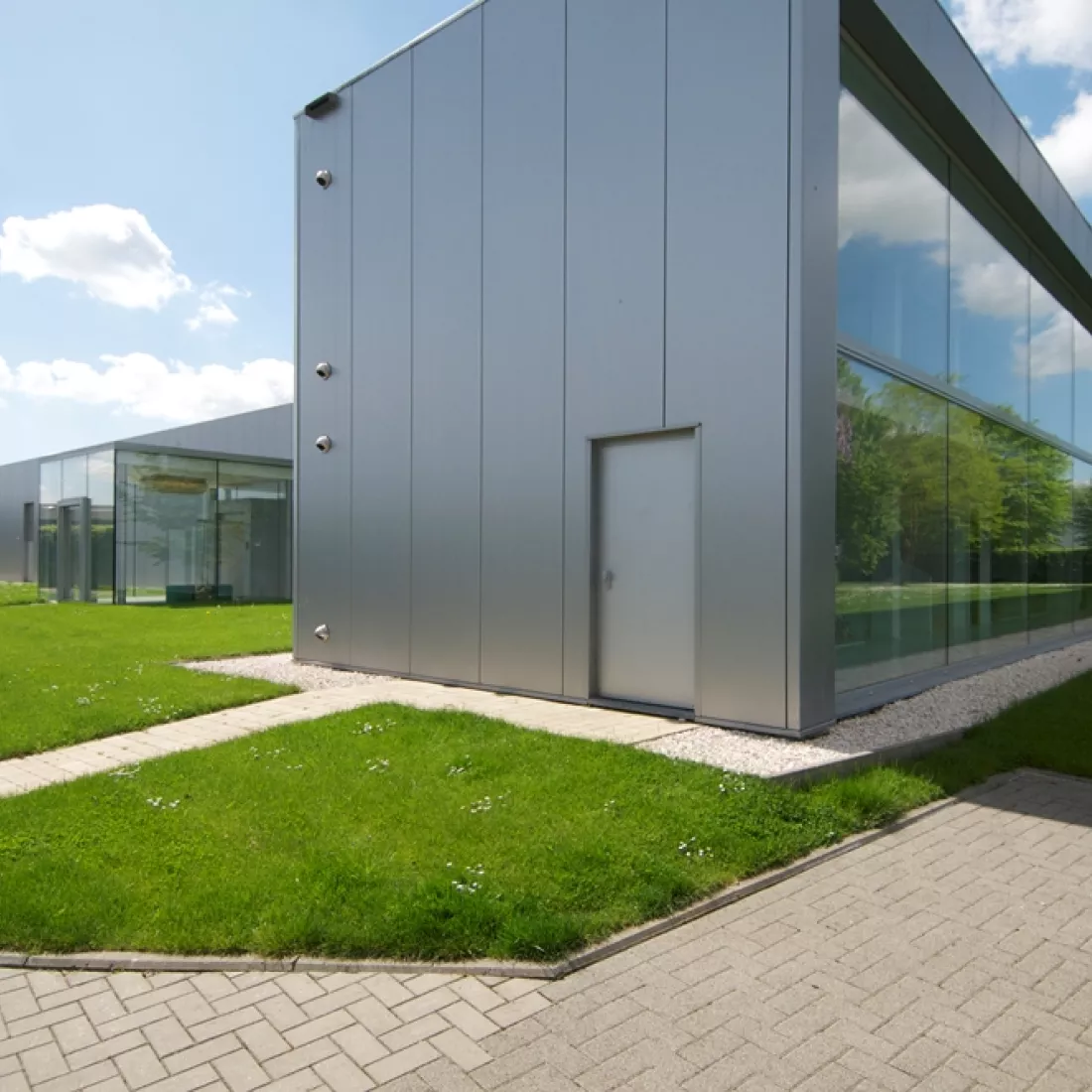 Zijaanzicht kantoor JNL. Het gebouw is grijs en bevat veel ramen. Rond het bedrijf is veel gras.