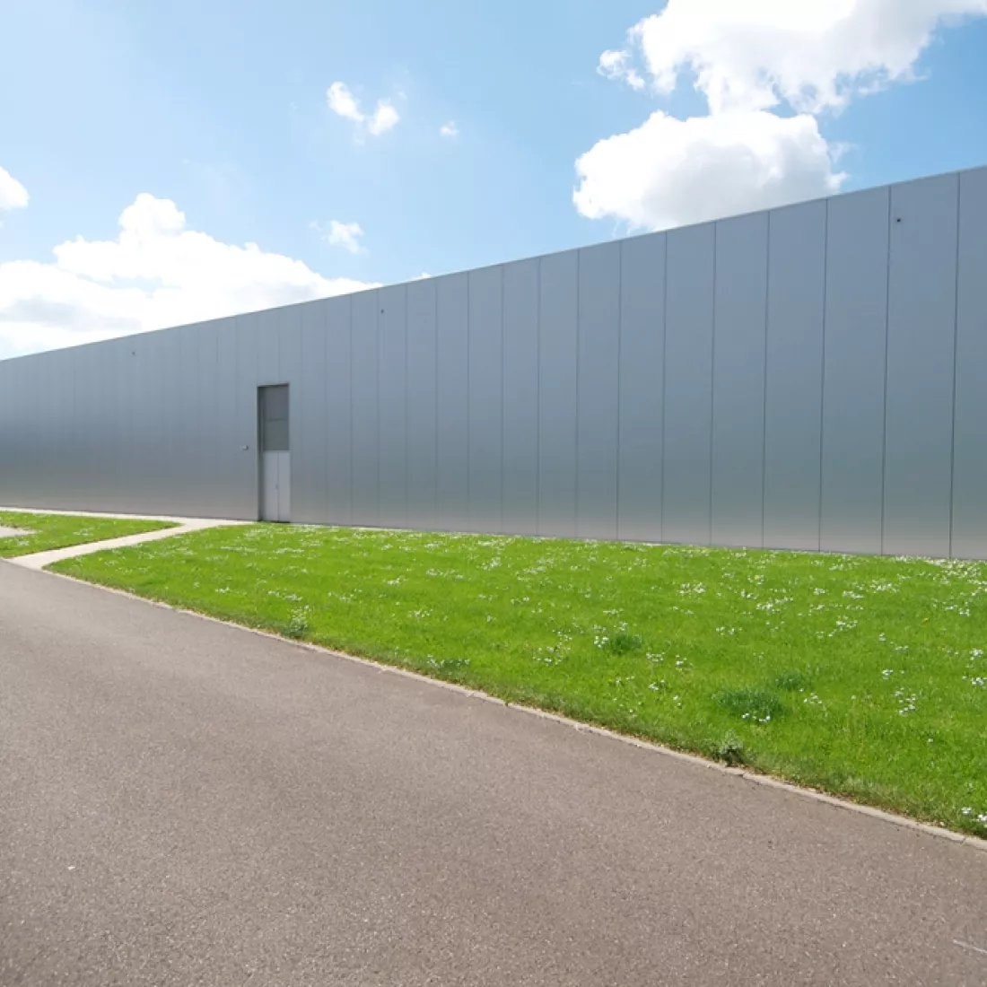 Buitenkant kantoor JNL. Dit deel van het gebouw bevat geen ramen; Het is een grijs gebouw. Ervoor is een grasveld en daarna een weg.