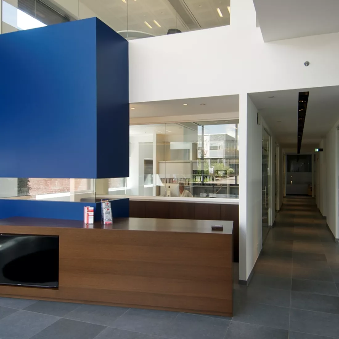 Binnenkant bedrijfspand Vitra. Rechts bevindt zich een gang en links het onthaal. Het onthaal bestaat uit een blauwe balie en een deel bestaat uit donkerbruin hout. Links bevindt zich een TV. Achter de balie bevindt zich een kantoor.