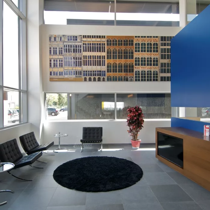 Binnenkant bedrijfspand Vitra. Rechts bevindt zich de balie. De balie bestaat uit donkerbruin hout en blauw. Er staan zwarte stoelen en er ligt een zwarte mat. De buitenmuren bevatten enkele ramen.