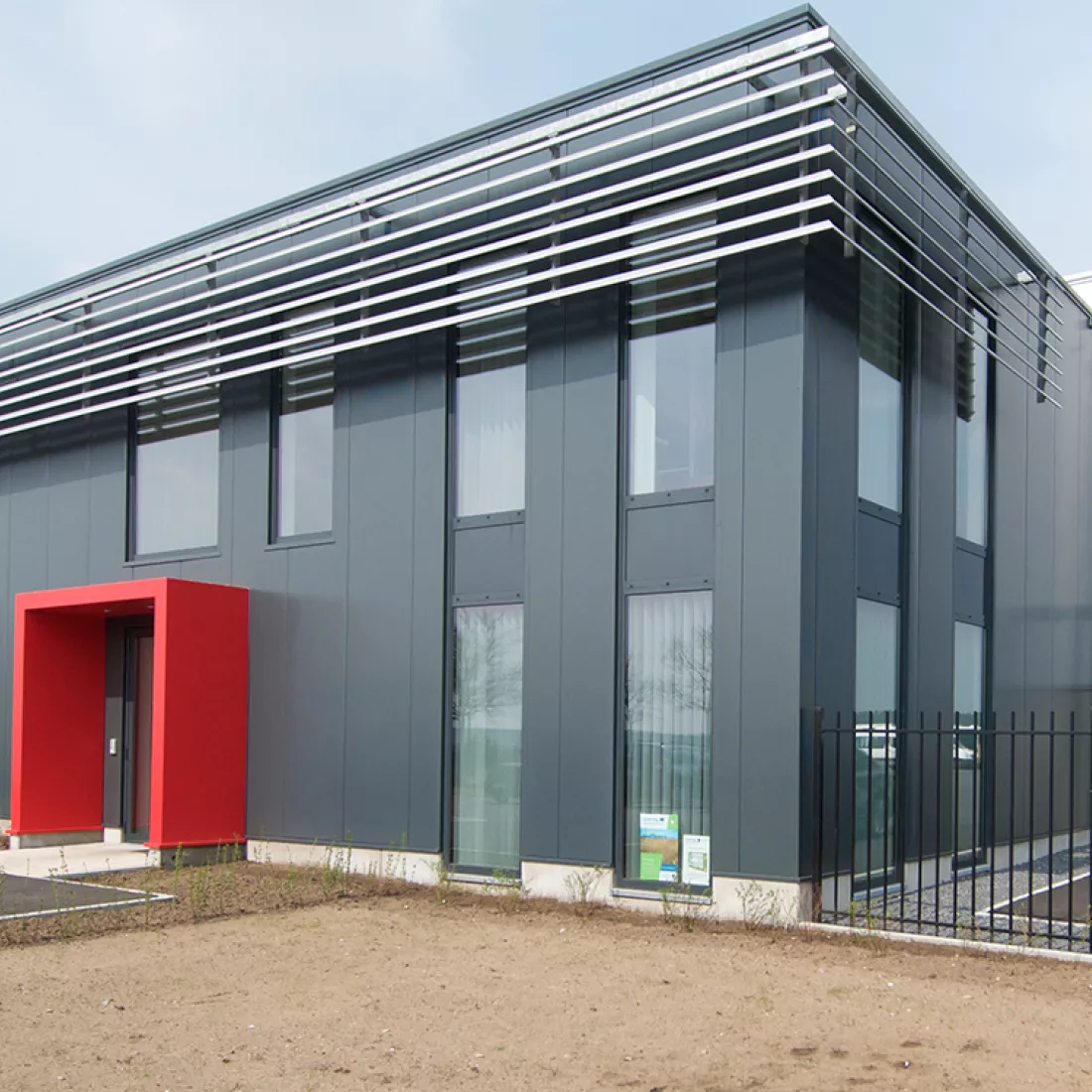 Zijaanzicht kantoor Hansea. Het gebouw is zwart en heeft rond de deur een rood detail. Het kantoor bevat ook veel smalle ramen. Rechts naast het kantoor is een zwart hek.