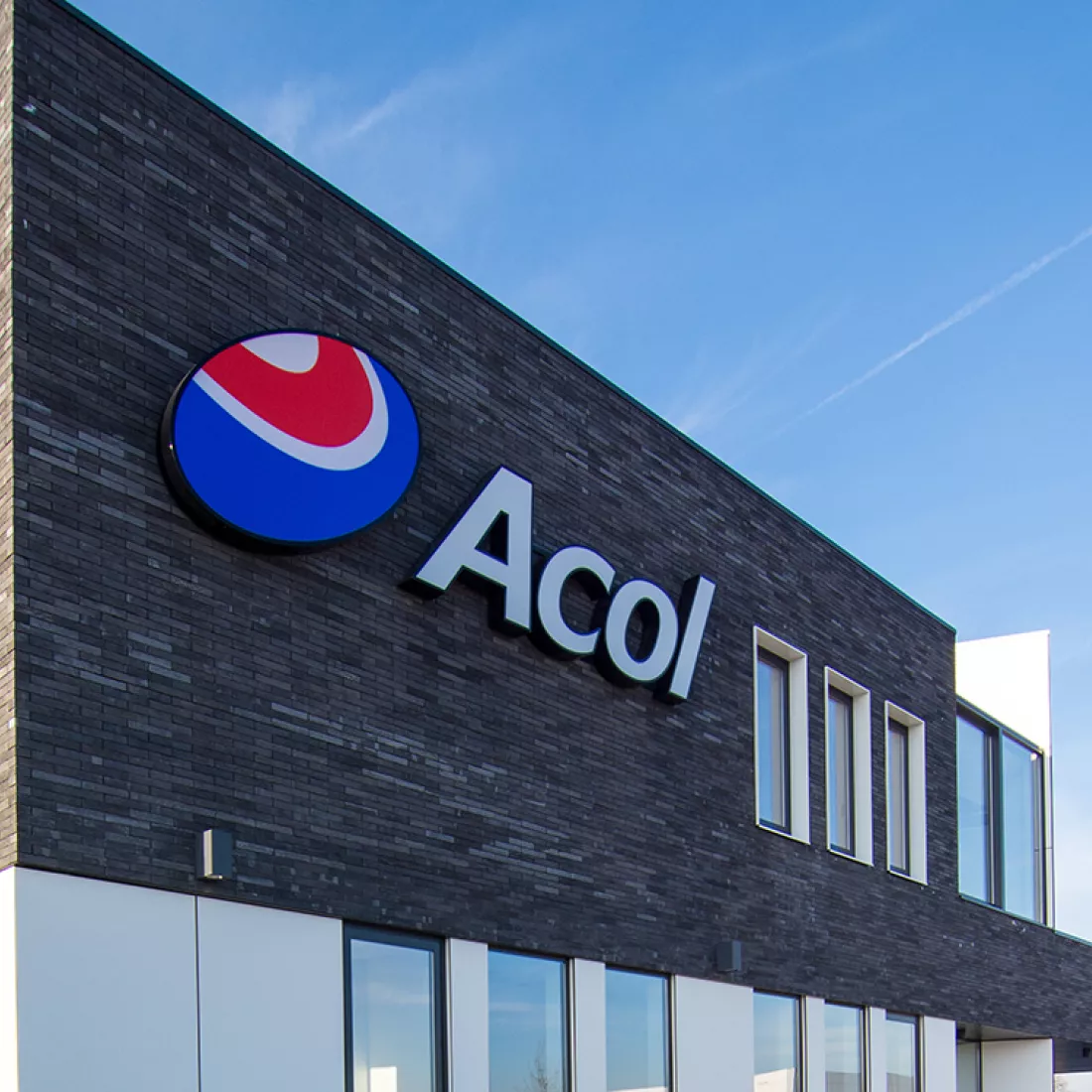 Close-up buitenkant kantoor Acol. De eerste verdieping bestaat uit zwarte steen met links het logo van Acol. De gelijkvloers is wit. Het kantoor bevat veel ramen.
