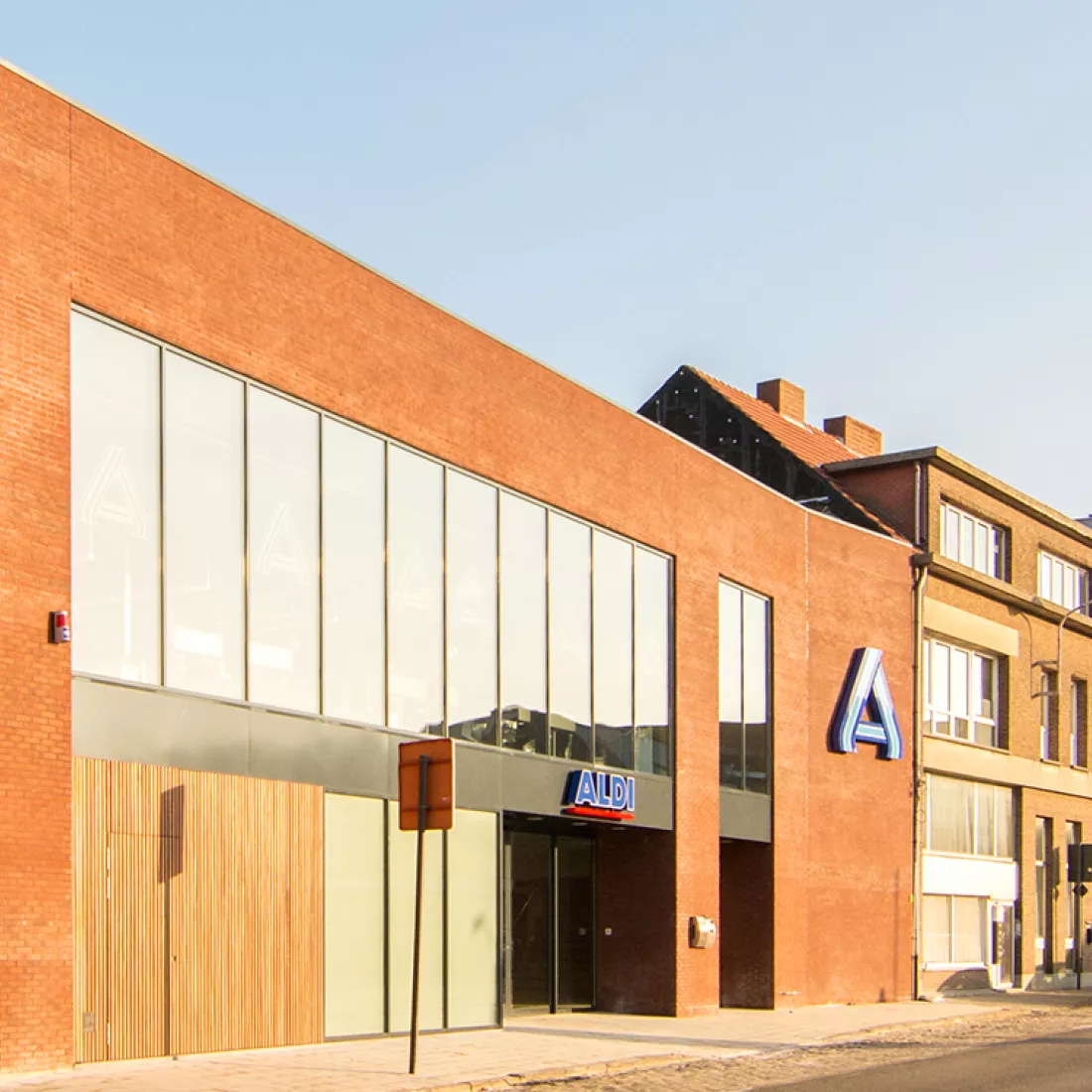 Vooraanzicht Aldi. De winkel is opgebouwd met een combinatie van materialen. Het gebouw bestaat uit een rode steen. Rechts hangt het logo tegen de muur. De winkel bevat grote ramen.