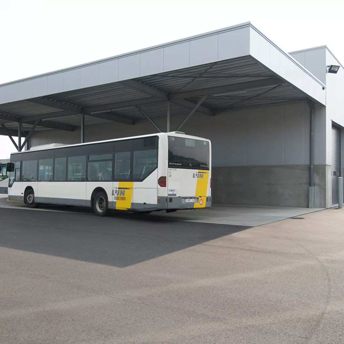 Zijaanzicht magazijn Hansea. Voor het magazijn staat een bus van De Lijn. Deze staat onder een afdak dat aan het magazijn hangt. Het magazijn is grijs en gemaakt met staal.