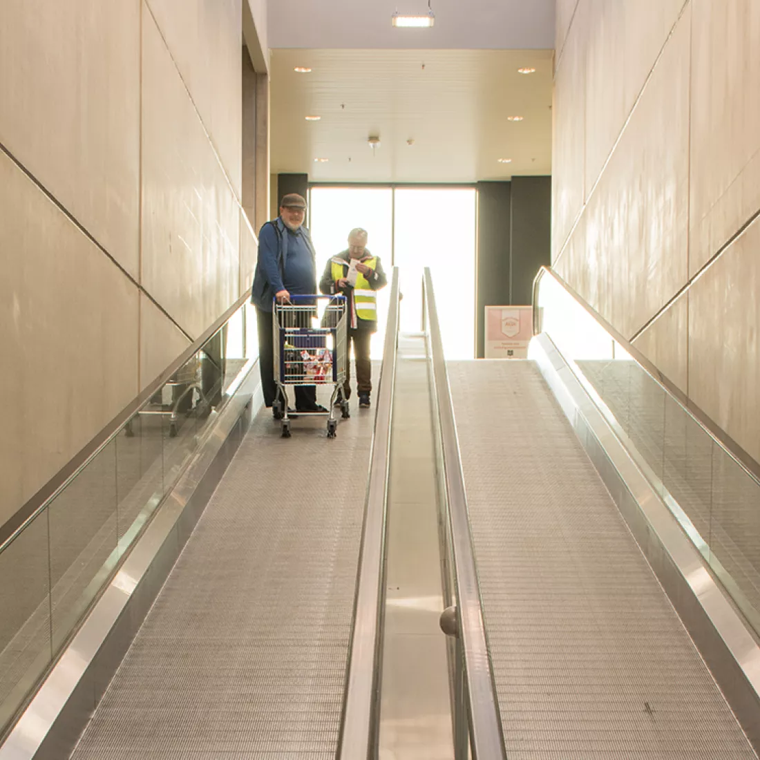 Binnenkant Aldi - rolpaden. Het linker rolpad gaat naar beneden en het rechter naar boven. Achteraan op de tweede verdieping bevindt zich een groot raam.