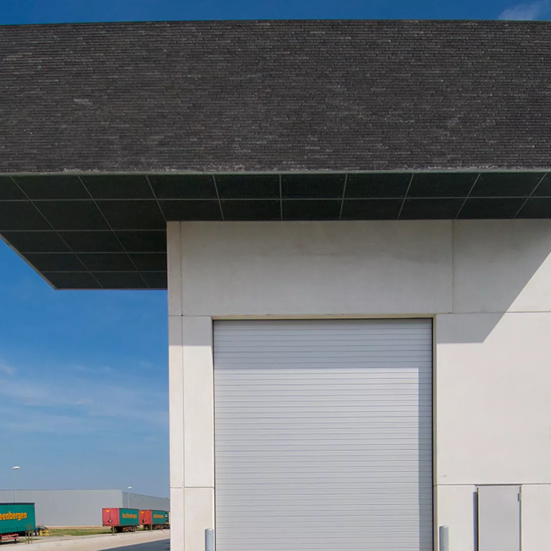 Vooraanzicht grijze poort productiehal Alcom. Boven de poort komt het detail van de zwarte steen van het kantoor terug.