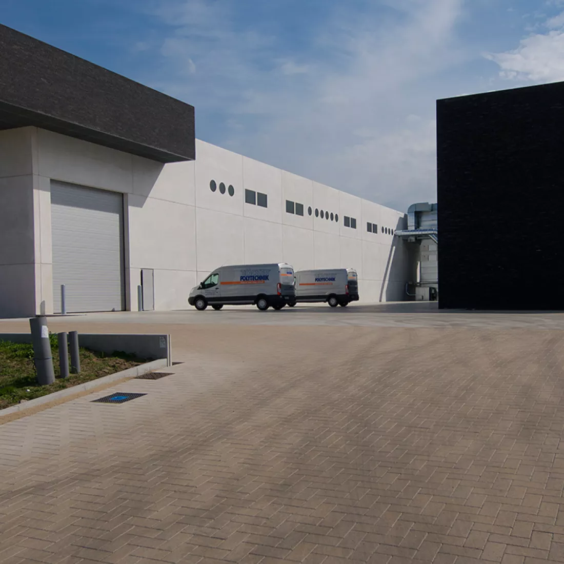 Productiehal Alcom. De productiehal is grijs met een grijze poort. Aan de linker bovenkant komt er een detail van de zwarte steen van het kantoor terug.